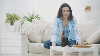 一个年轻的女人坐在沙发上，吃披萨，喝啤酒。 她正在打开电视。 好吃-好吃。 4K.